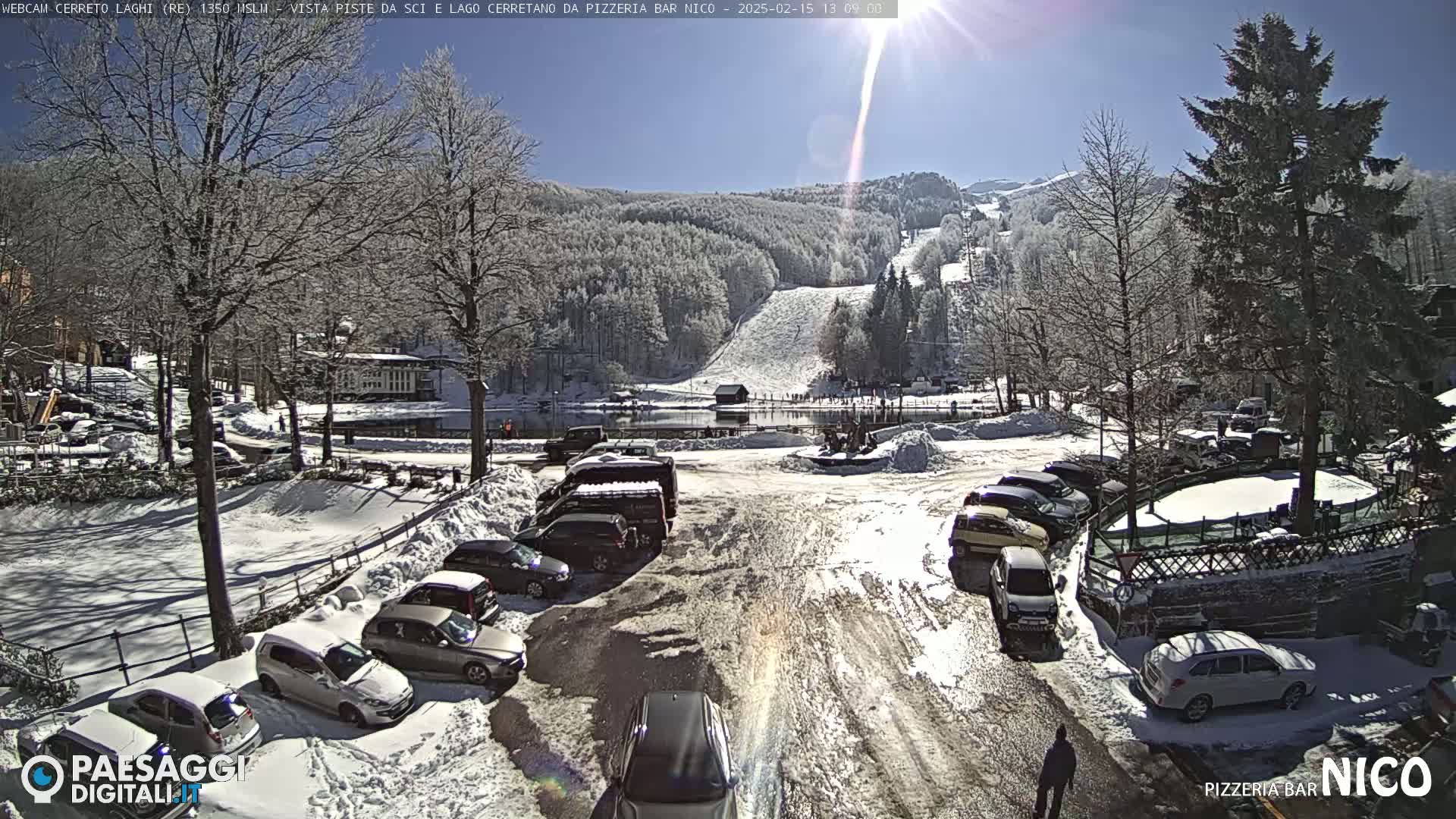 A sunny, snow-covered parking lot with cars parked near a frozen lake and ski slope in the background.