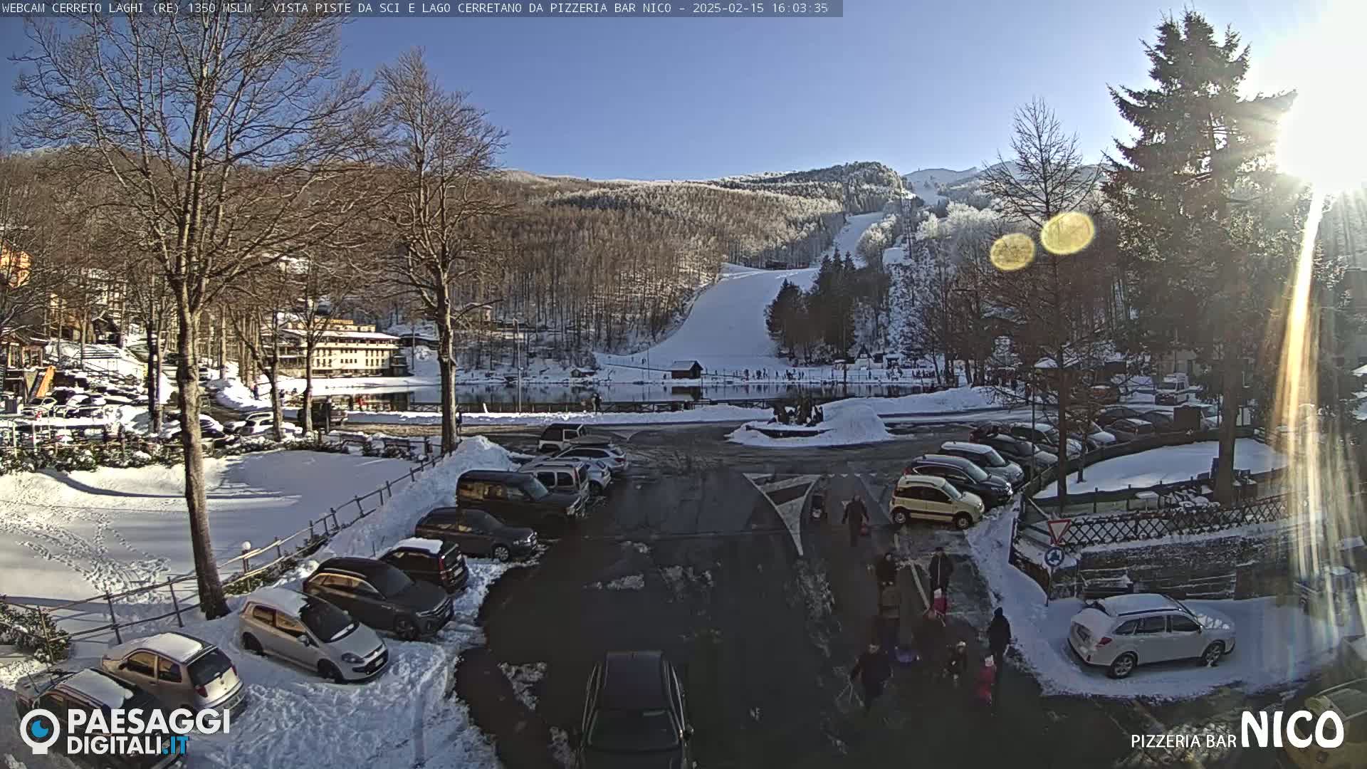 A sunny, snow-covered parking lot with cars and people, situated near a frozen lake and ski slopes in a mountainous area.