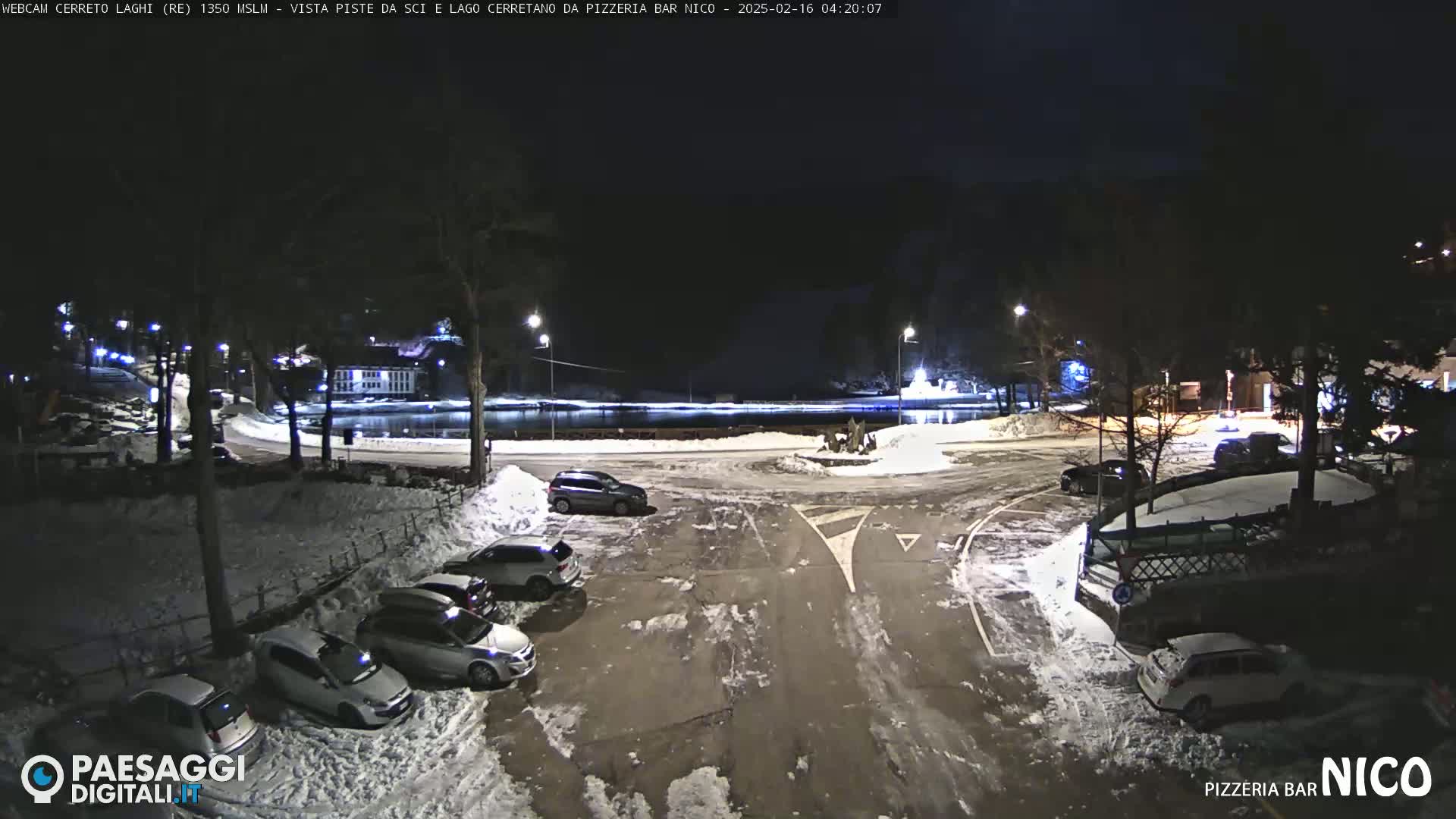 A snow-covered parking lot at night with several cars parked, illuminated by streetlights, near a body of water.