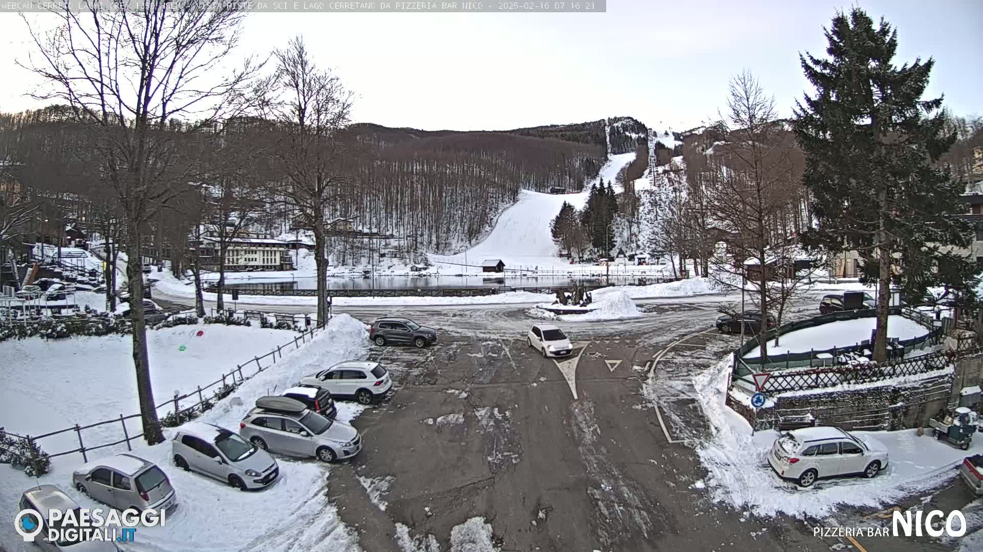 A snowy parking lot with several cars is situated in front of a small town nestled at the base of a snow-covered hill with a ski slope.