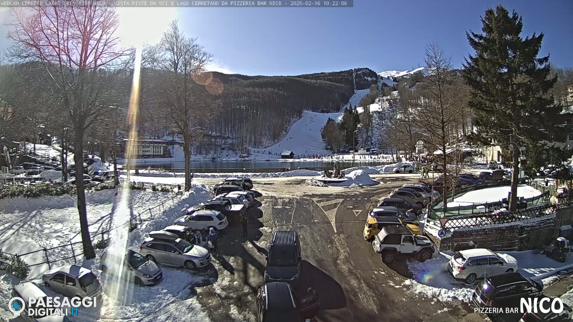 A sunny winter day shows a parking lot full of cars near a frozen lake and a ski slope in the background.