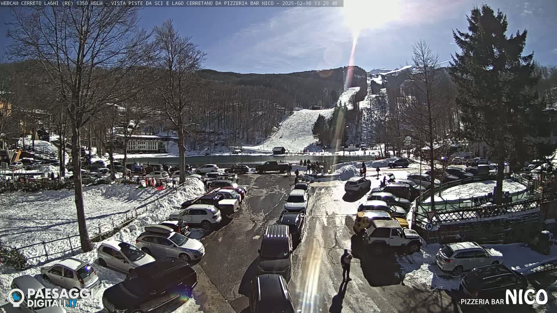 A sunny, snowy parking lot is full of cars, with a ski slope and snow-covered mountain visible in the background.