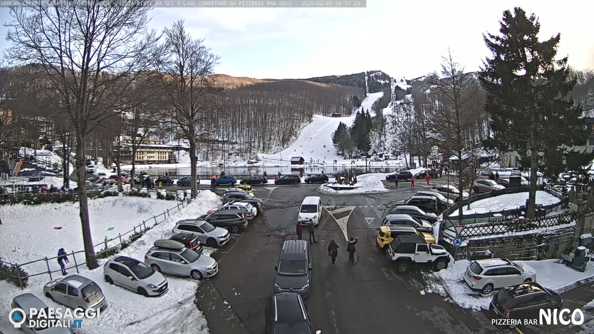 A snowy parking lot with cars and people surrounds a frozen lake, with a ski slope visible in the background under a partly cloudy sky.
