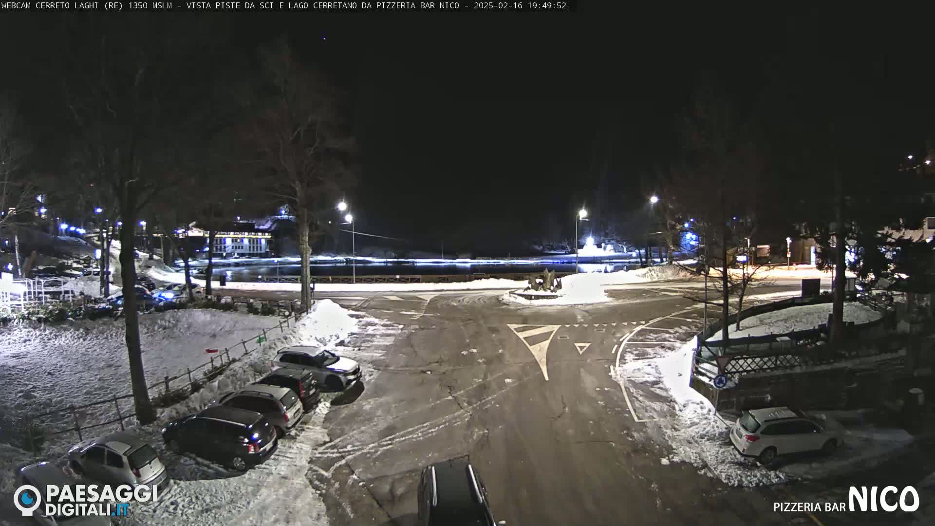 A snow-covered parking lot at night, illuminated by streetlights, with several cars parked and a road leading to a roundabout near a body of water.