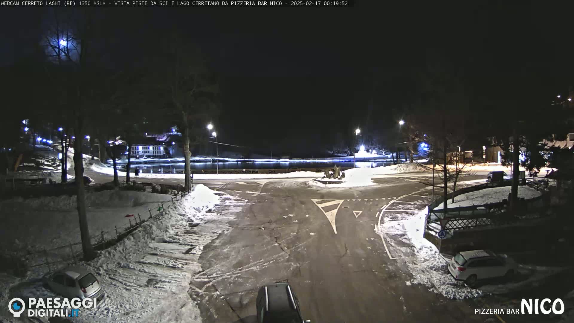 A nighttime, snow-covered parking lot and roadway with several cars and a small body of water visible in the background under a mostly clear sky.