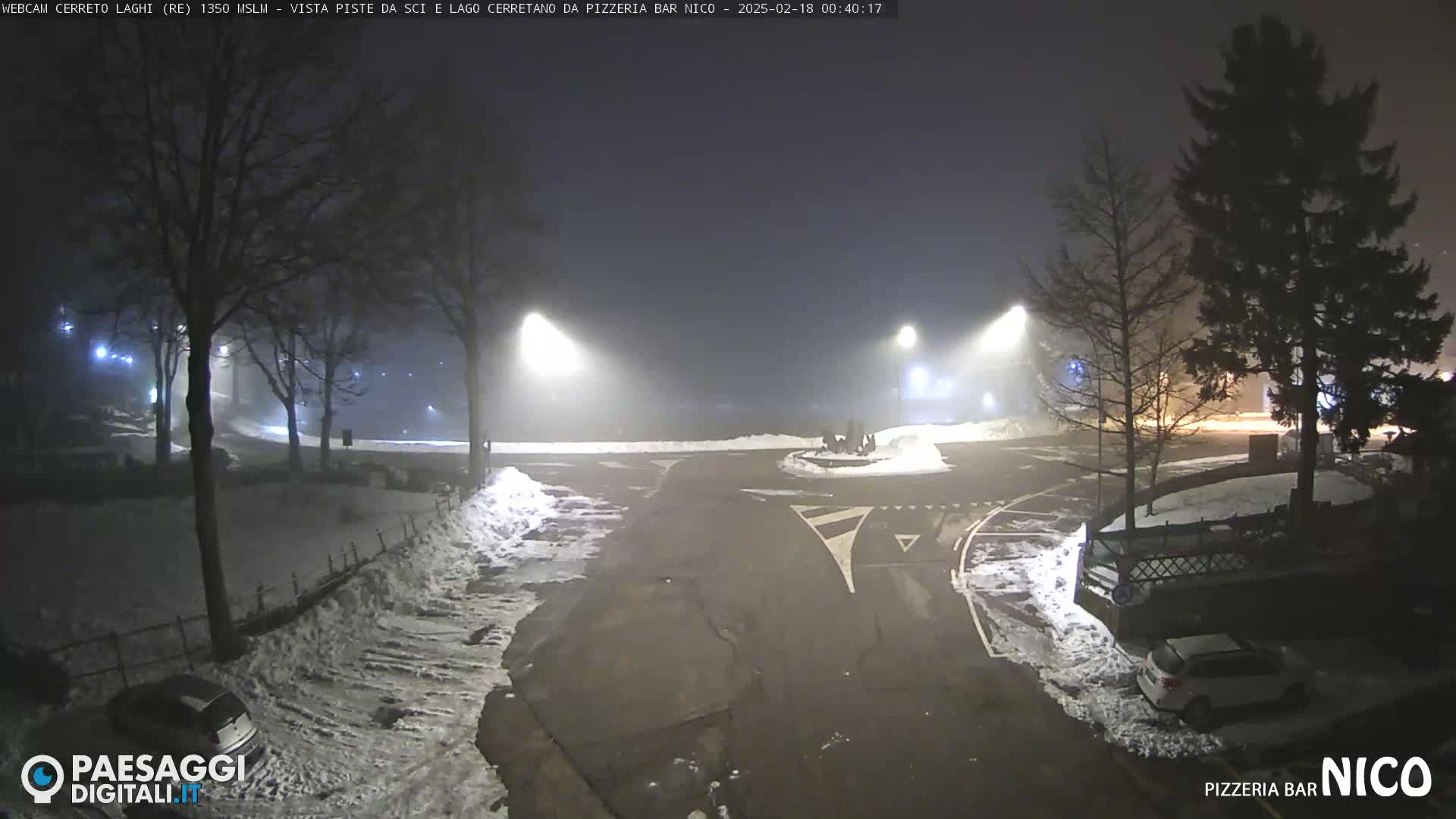 A snow-covered nighttime scene shows a roadway intersection with several cars parked, illuminated by streetlights in a foggy atmosphere.