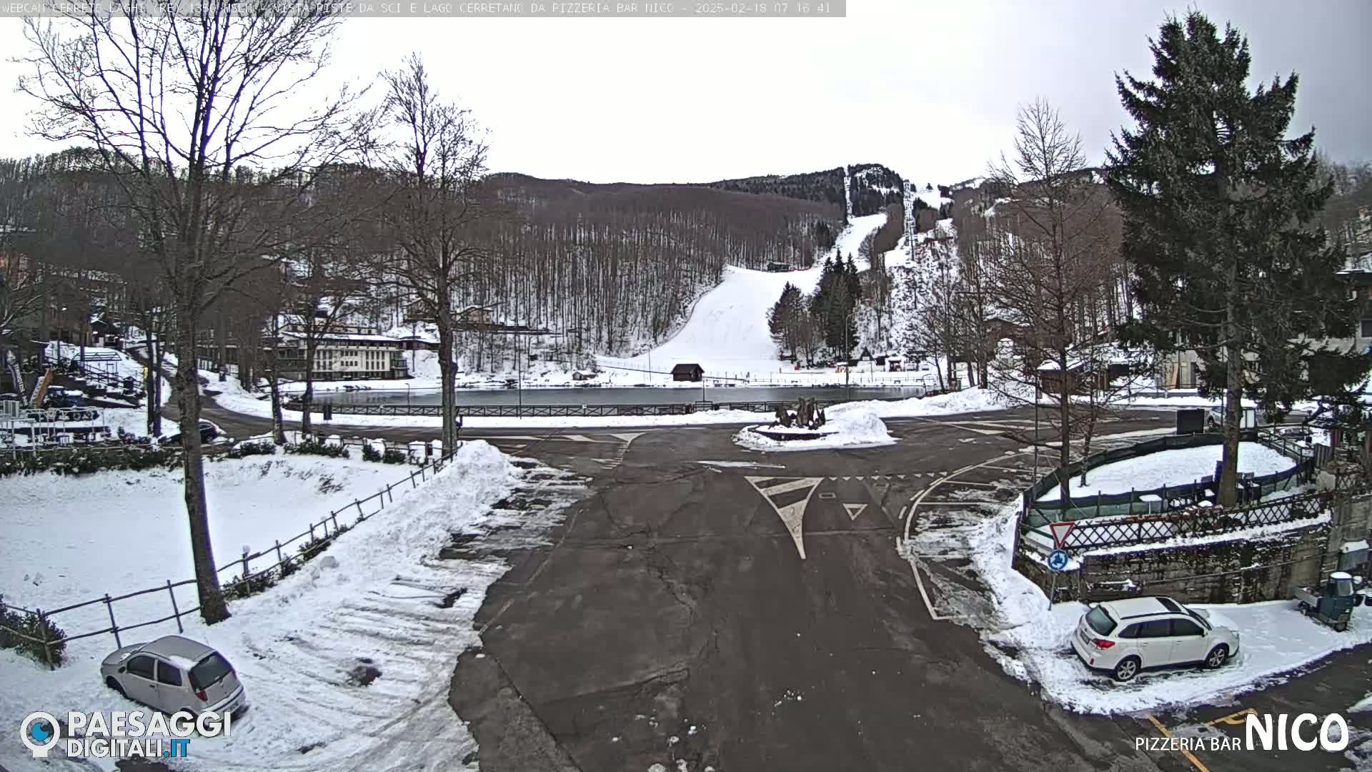 A snowy parking lot and roadway scene overlooks a frozen pond and a ski slope on a cloudy day.