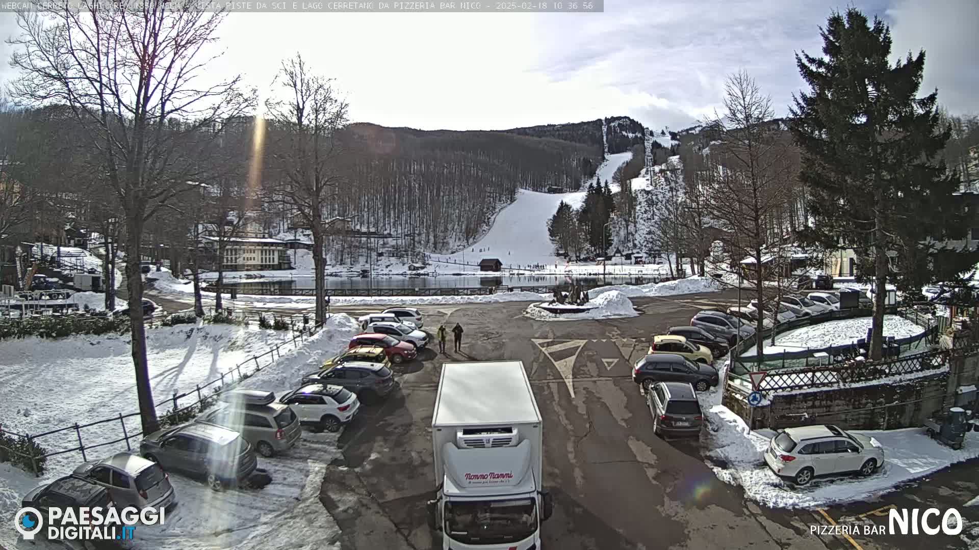 A sunny winter scene shows a parking lot filled with cars next to a frozen lake, with a ski slope and snow-covered trees in the background.
