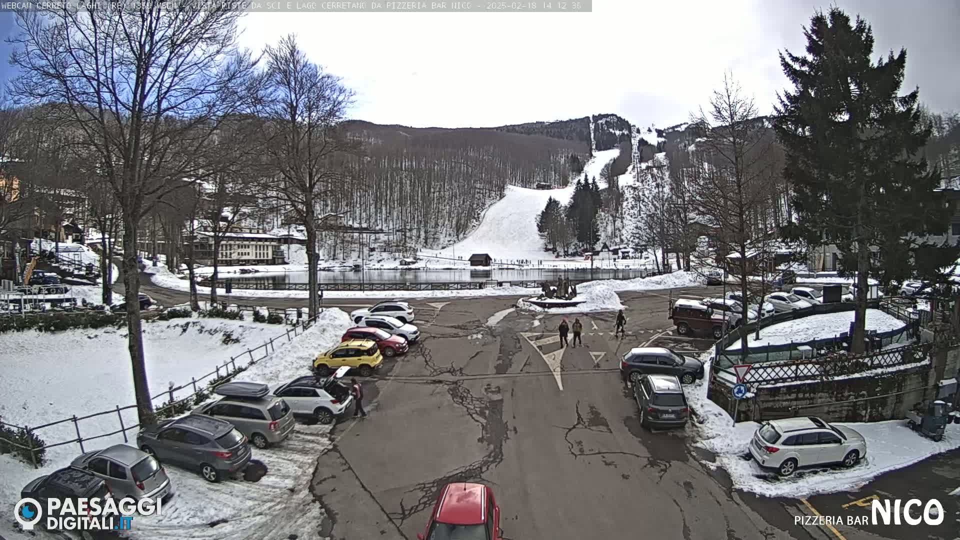 A snow-covered parking lot with cars and a few people walking, overlooks a ski slope and a frozen lake, under a mostly cloudy sky.