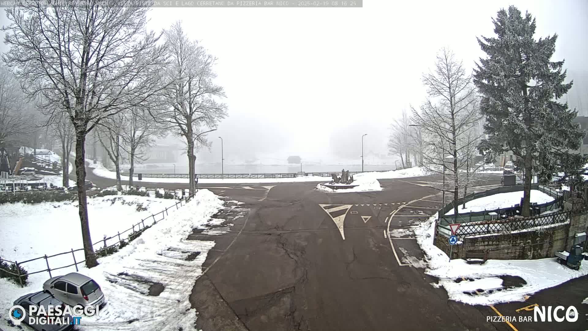 A snow-covered parking lot and roadway intersect near a lake in foggy conditions, with bare, snow-dusted trees surrounding the area.
