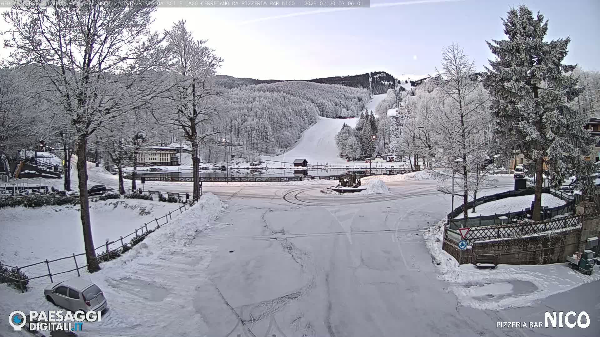 A snow-covered parking lot and roundabout are situated in front of a snow-dusted ski slope and forested mountain, under a clear, light sky.