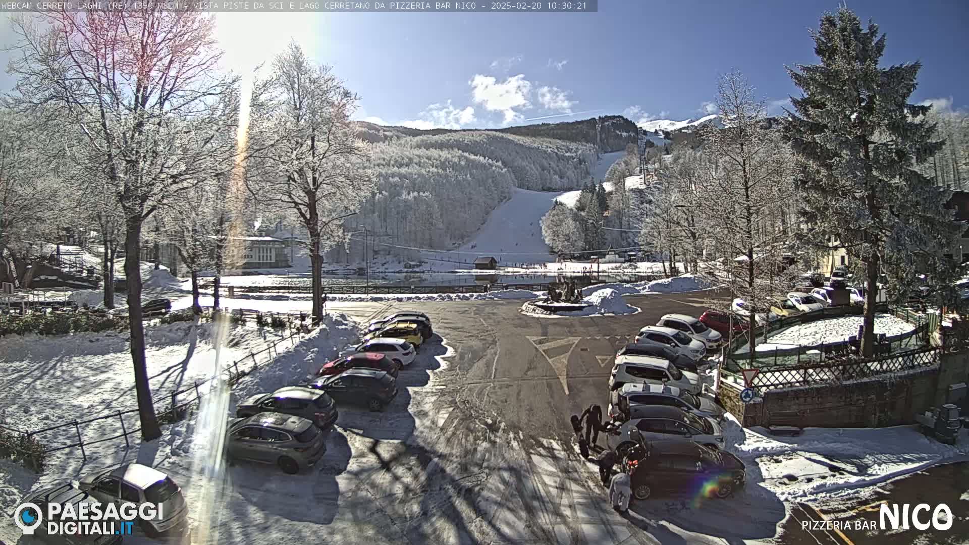 A sunny, snow-covered parking lot with cars and a background showing a ski slope on a mountainside.