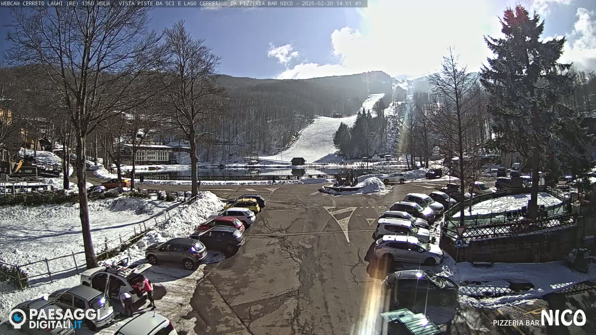 A sunny, snow-covered parking lot with cars and a view of a ski slope and a lake in the background.