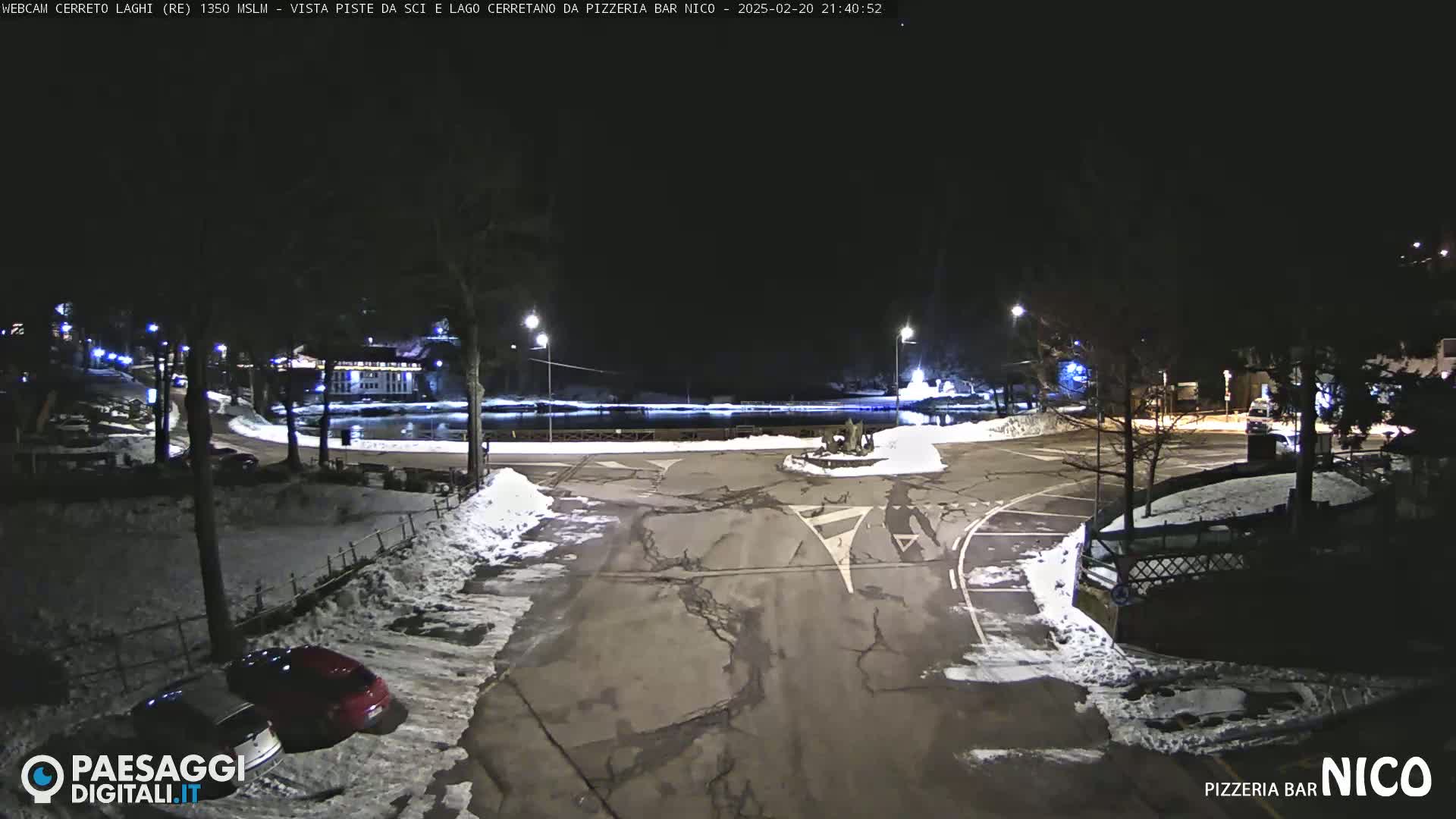 A nighttime view of a snow-covered parking lot and a frozen lake, illuminated by streetlights.