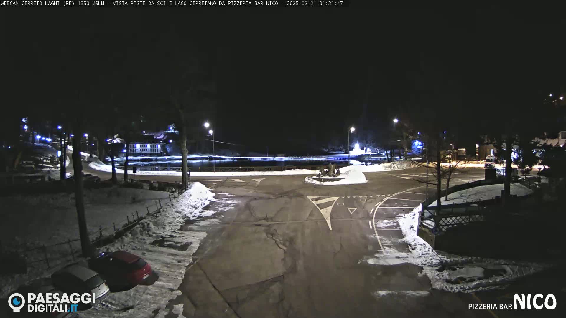 A snow-covered parking lot and roundabout are illuminated at night near a body of water.