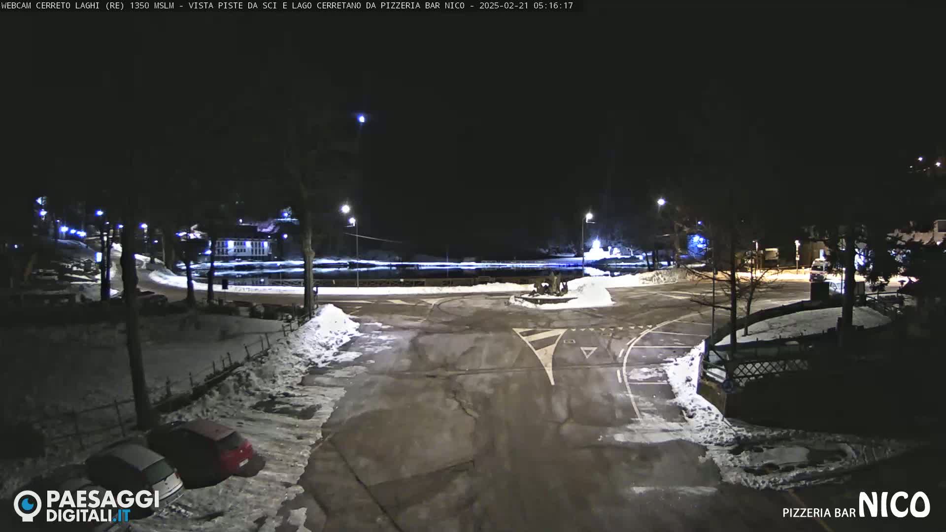 A snow-covered parking lot and roadway are illuminated at night, with several cars parked and a few bare trees visible.