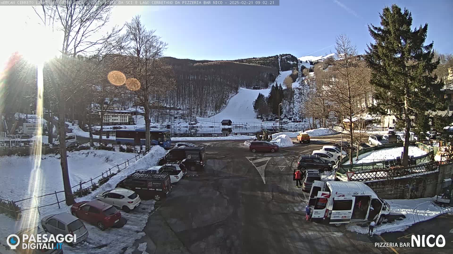 A sunny winter day shows a parking lot with several cars and vans, next to a snow-covered ski slope and wooded hillside.