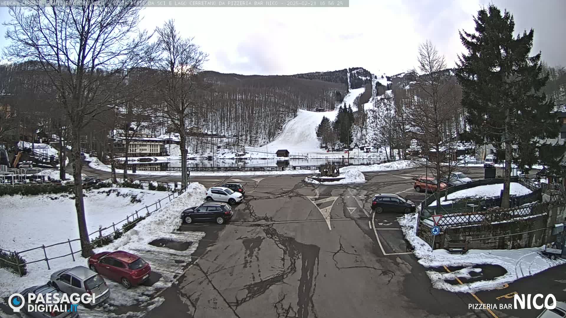 A snowy parking lot with several cars is situated in front of a building, overlooking a ski slope on a partly cloudy day.