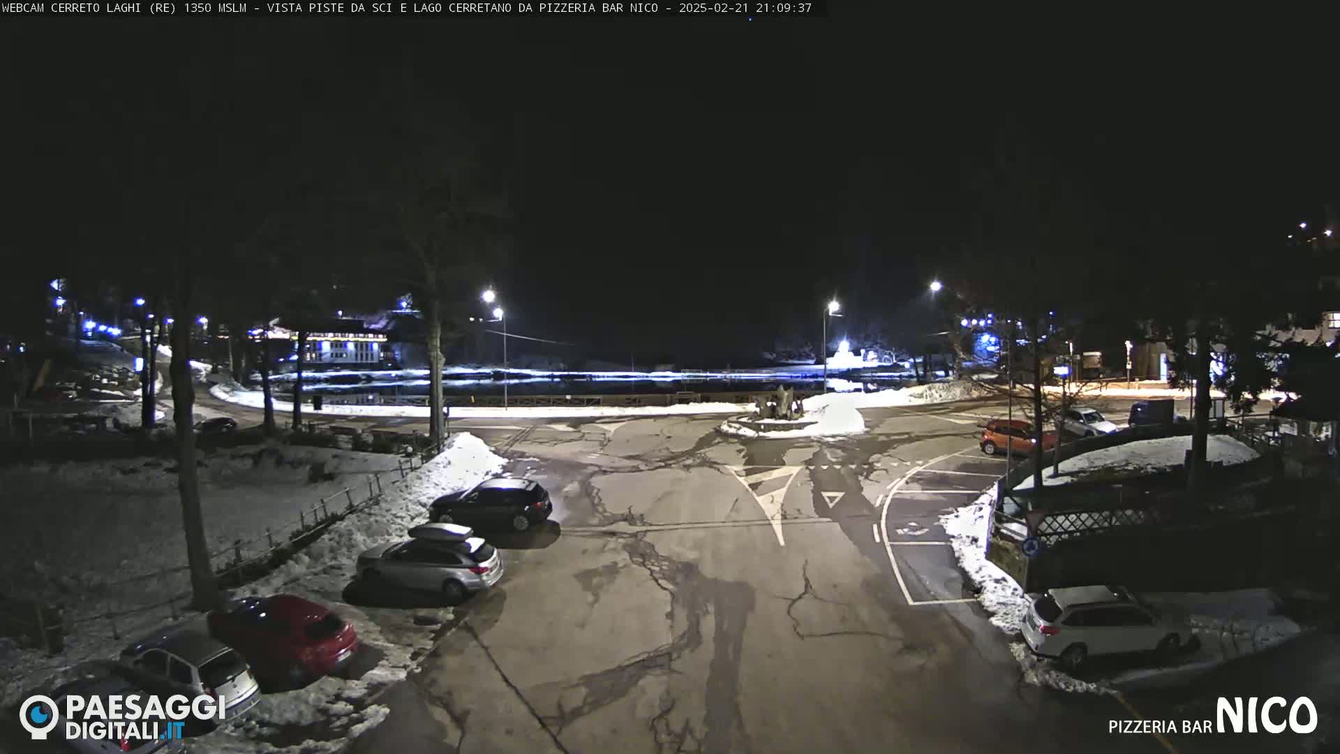 A nighttime, snow-dusted parking lot with several cars, surrounded by trees and buildings under dark skies.