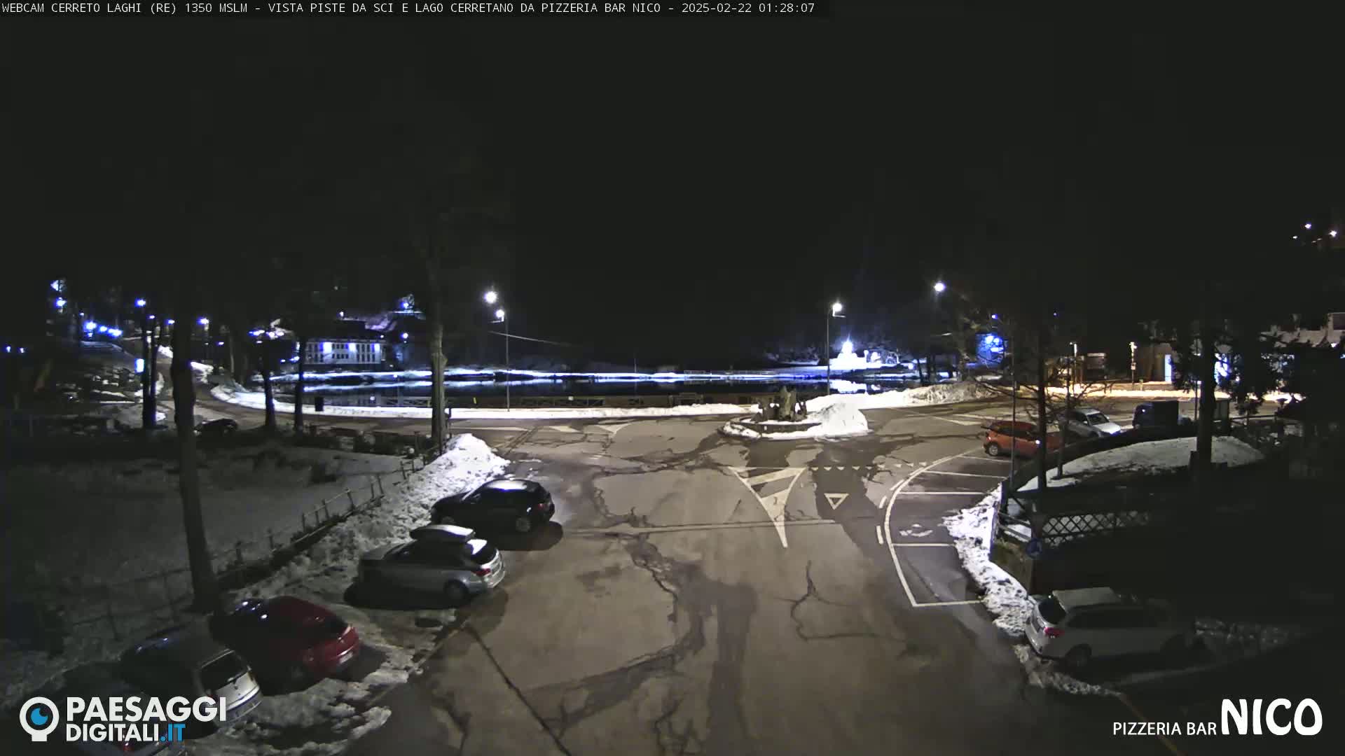 A nighttime view of a snow-dusted parking lot with several cars parked, situated near a body of water, under a clear, dark sky.