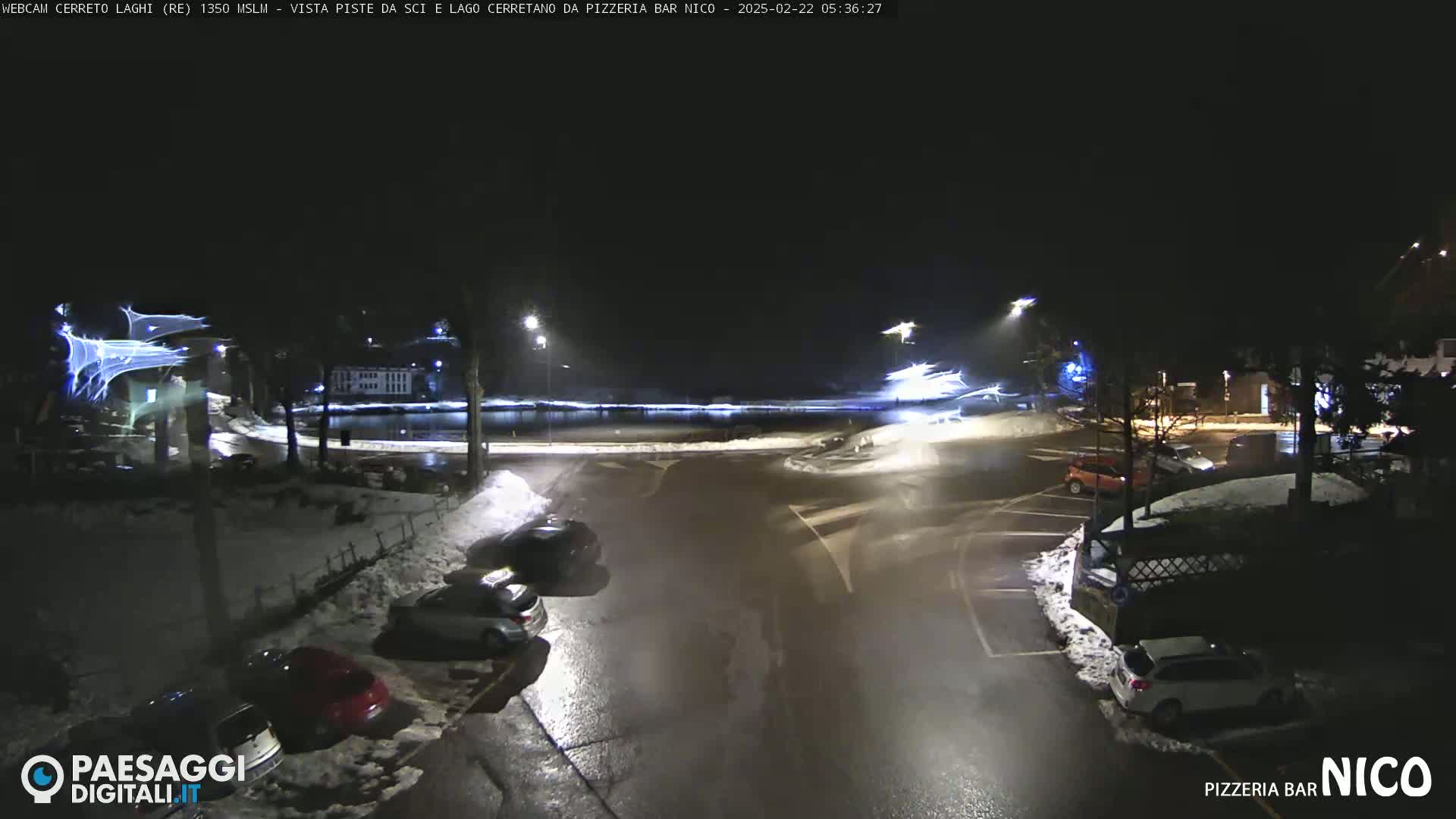 A nighttime scene of a snow-covered parking lot and road intersection with several cars parked, illuminated by streetlights, and faint streaks of light in the sky.