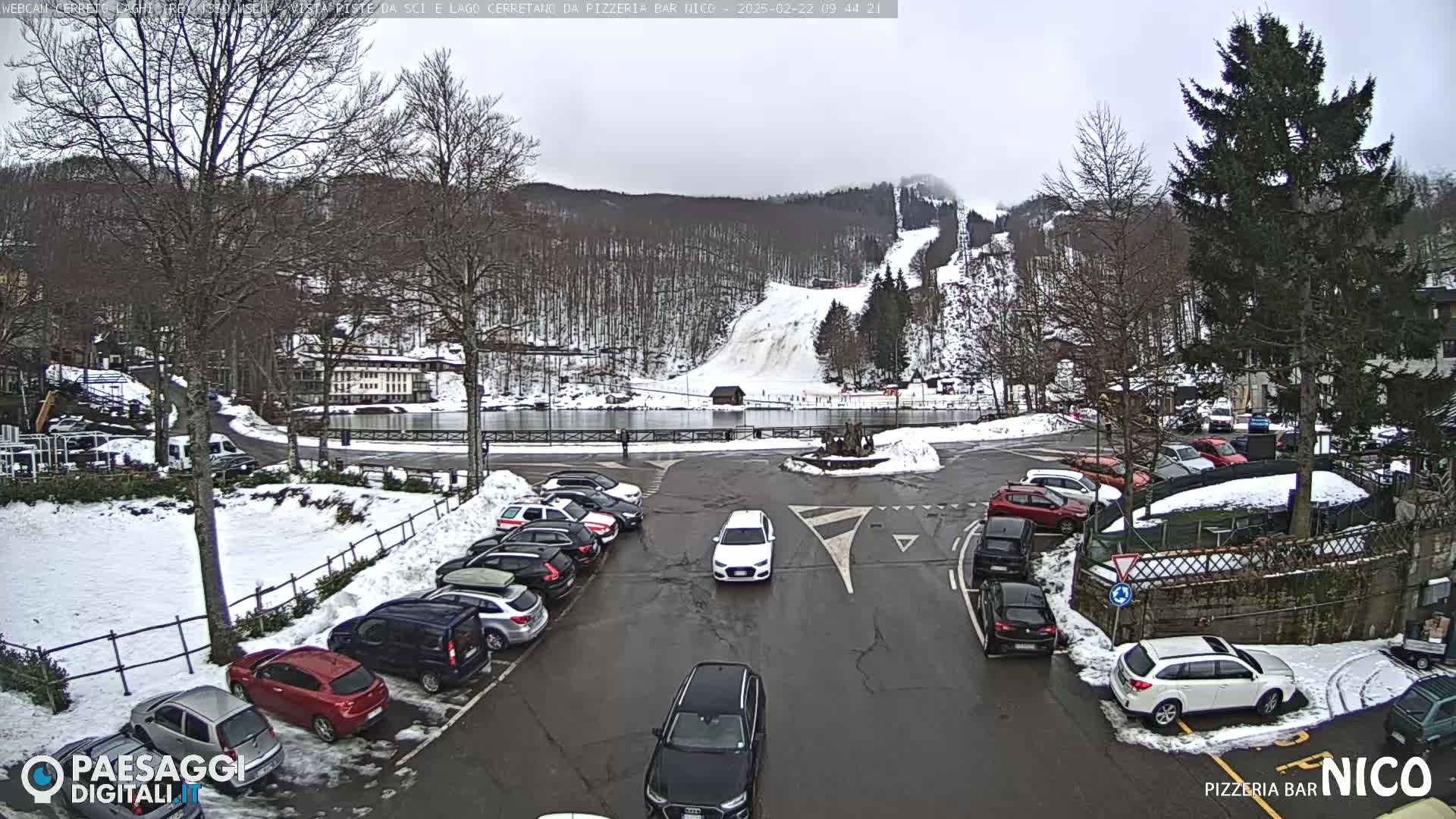 A parking lot with numerous cars is situated in front of a ski slope and a building, under an overcast sky with some snow on the ground.