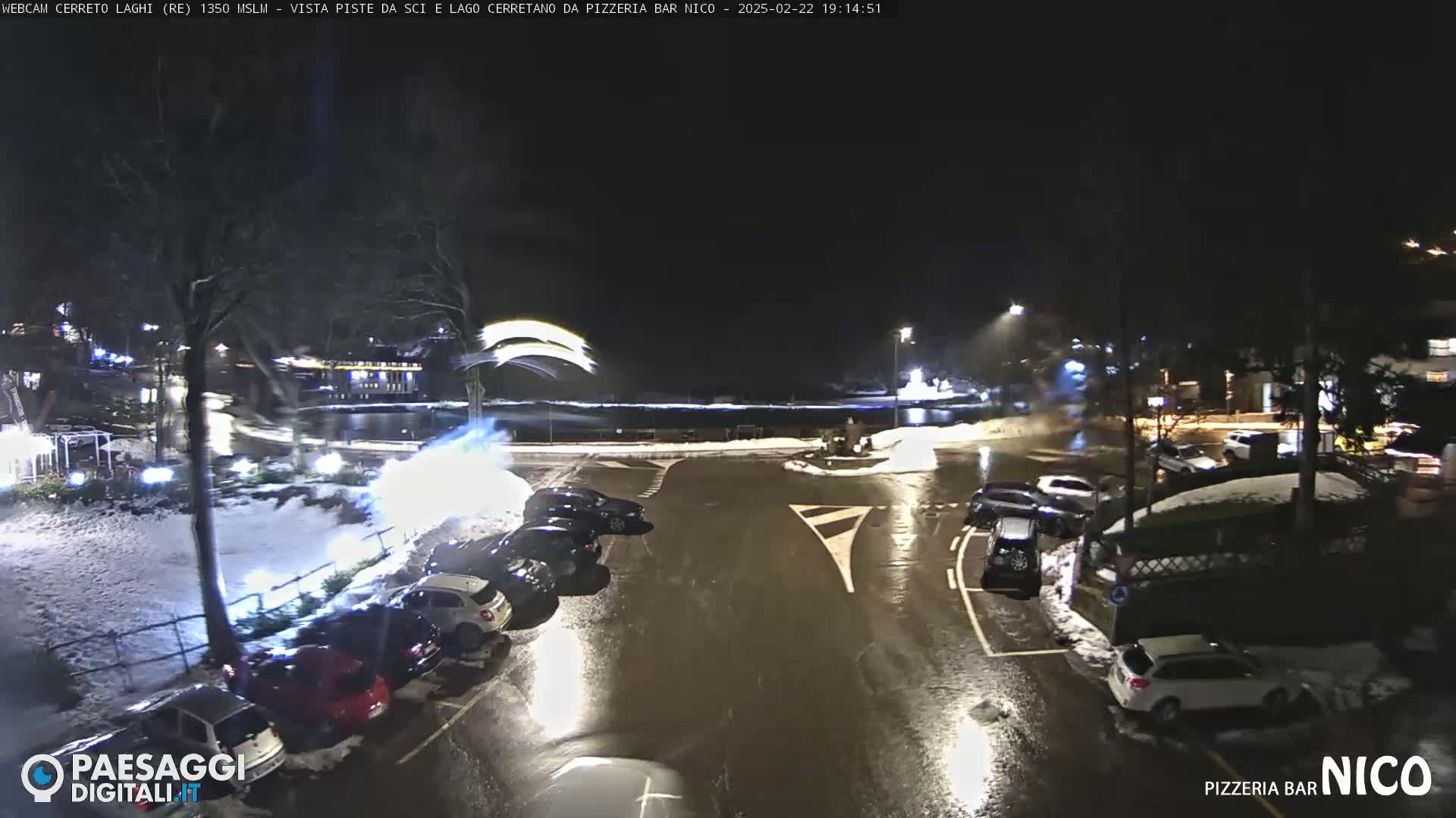 A nighttime parking lot with cars parked along the edge, wet pavement, and some snow on the ground, with faint artificial light in the distance.