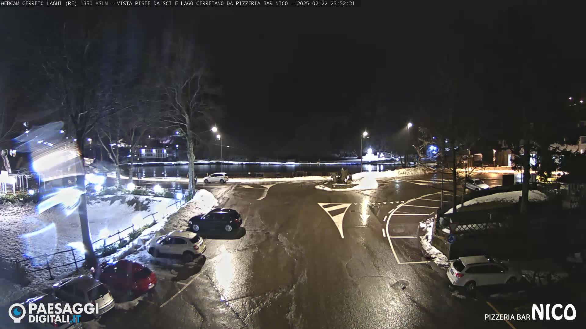 A nighttime view of a parking lot with several cars, partially covered in snow, next to a calm body of water under a dark sky.