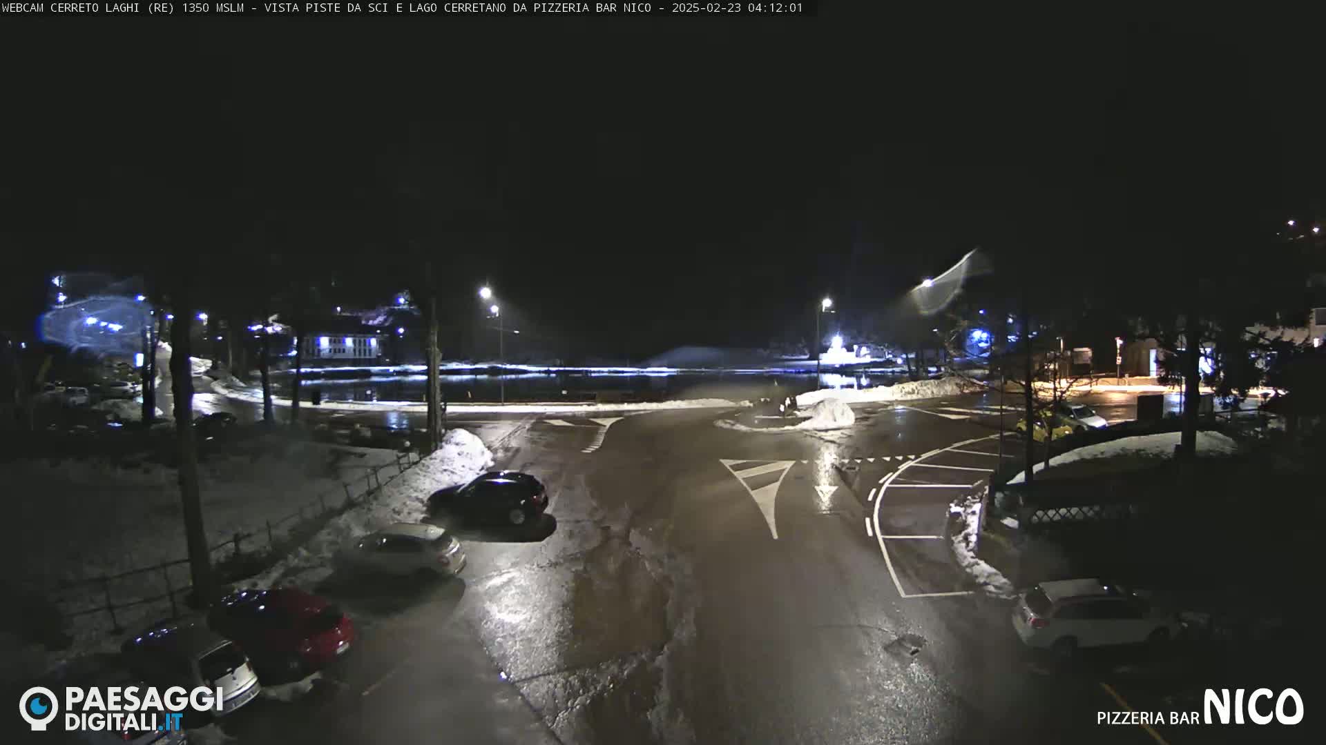 A nighttime view of a parking lot and roadway, partially covered in snow, with several cars parked and a body of water visible in the background.