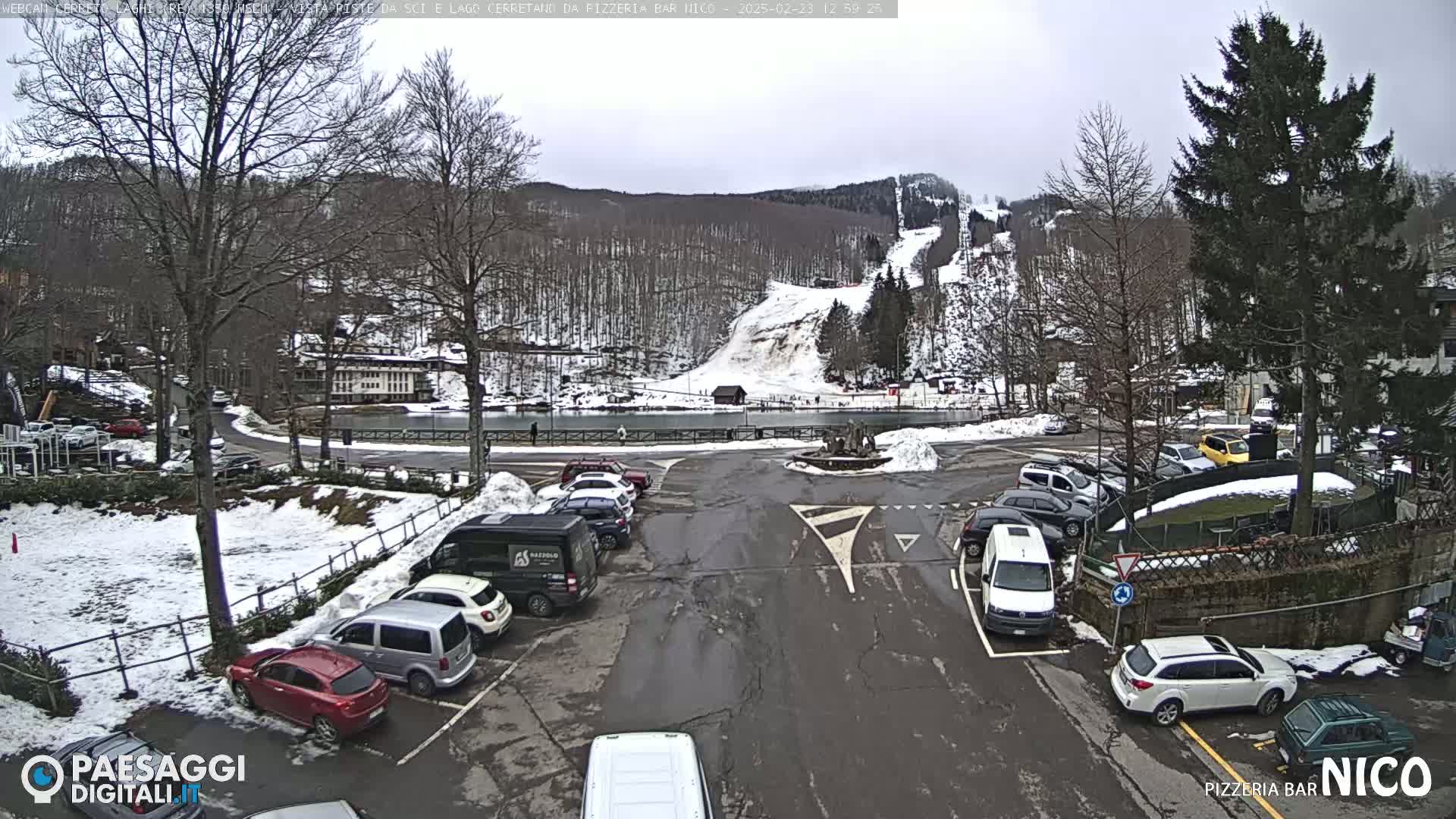 A parking lot with cars is situated in front of a building near a snowy ski slope and lake under an overcast sky.