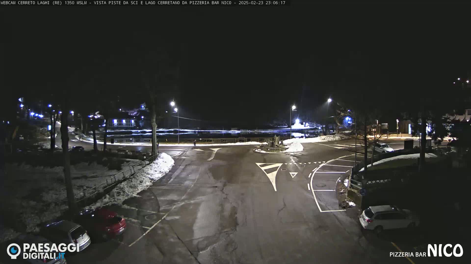 A nighttime view of a snow-dusted parking lot and roundabout, illuminated by streetlights.