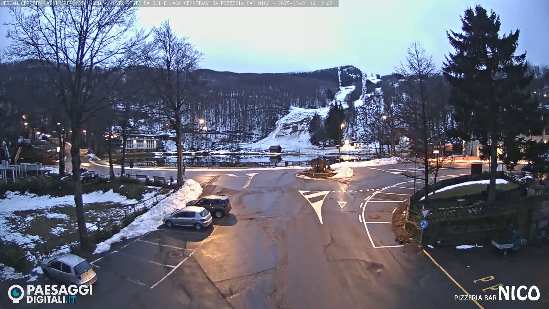 A parking lot with a few cars, a roundabout, and a snow-covered landscape with trees and ski slopes under a twilight sky.