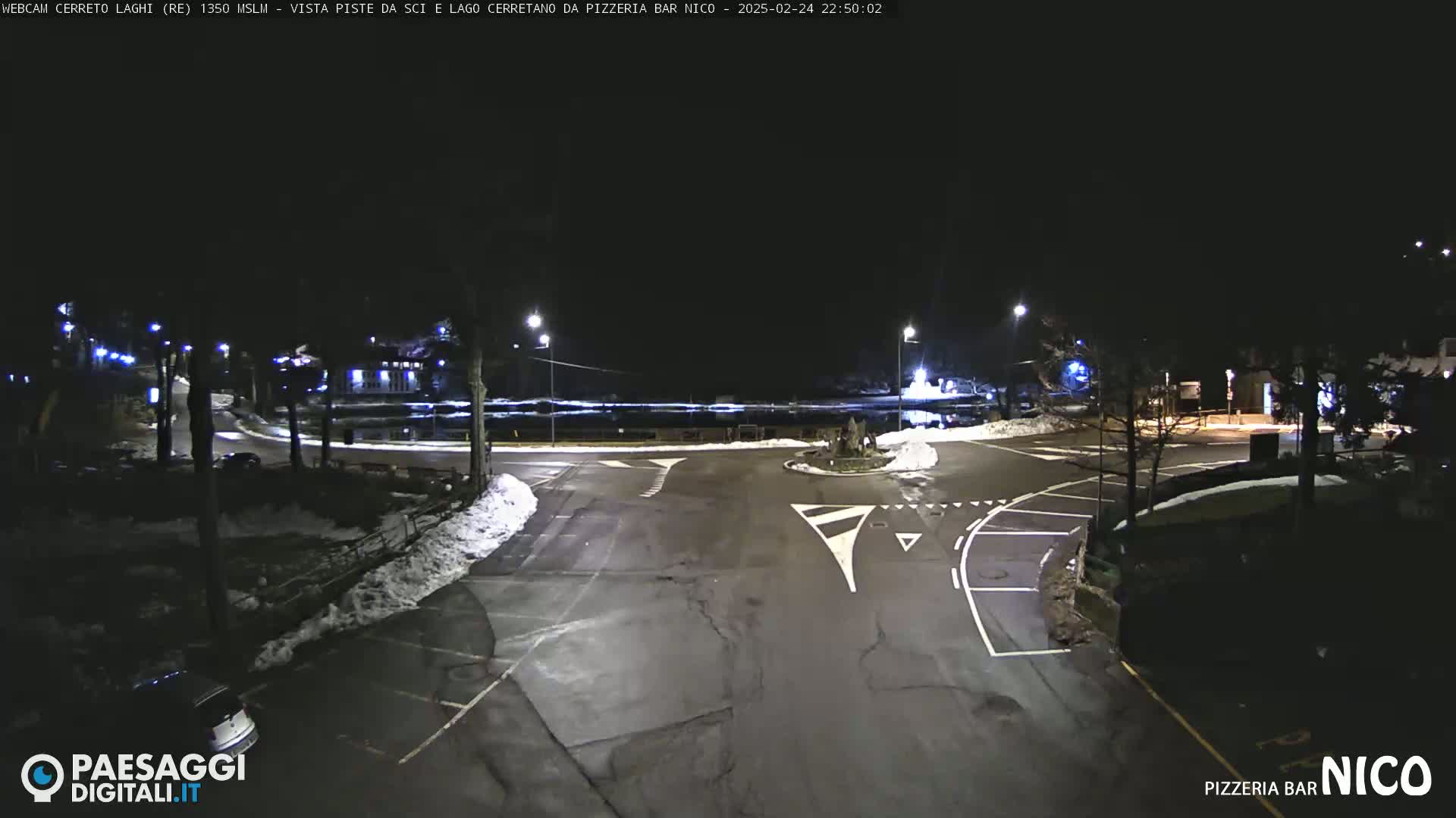 A dark, snow-dusted parking lot and roadway intersection are lit by streetlights at night.