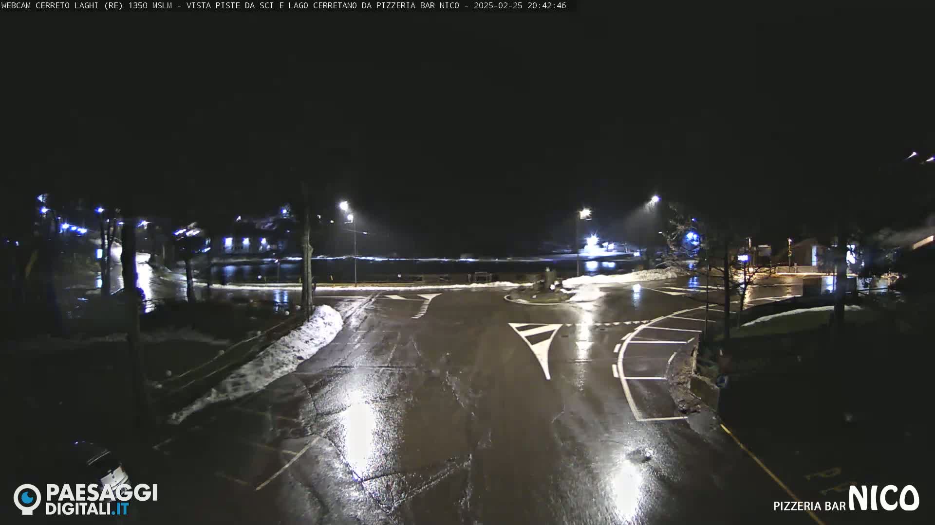 A wet, nighttime scene shows a parking lot and roadway intersection with some snow on the ground and streetlights illuminating the area.
