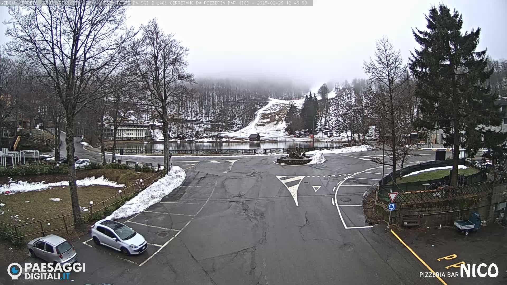 A mostly empty parking lot with some cars, surrounded by snow-covered ground and trees, overlooks a small lake and a snow-covered hillside under an overcast sky.