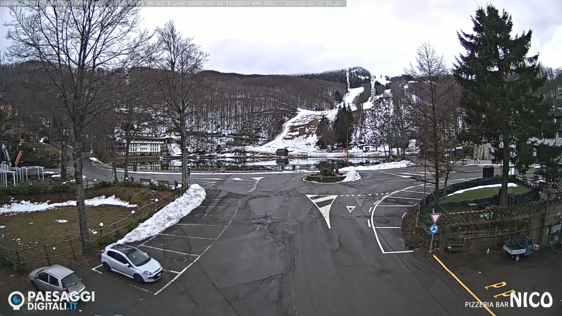 A parking lot with a few cars, partially covered in snow, overlooks a small lake and a snow-covered hillside with a ski slope in the background under an overcast sky.