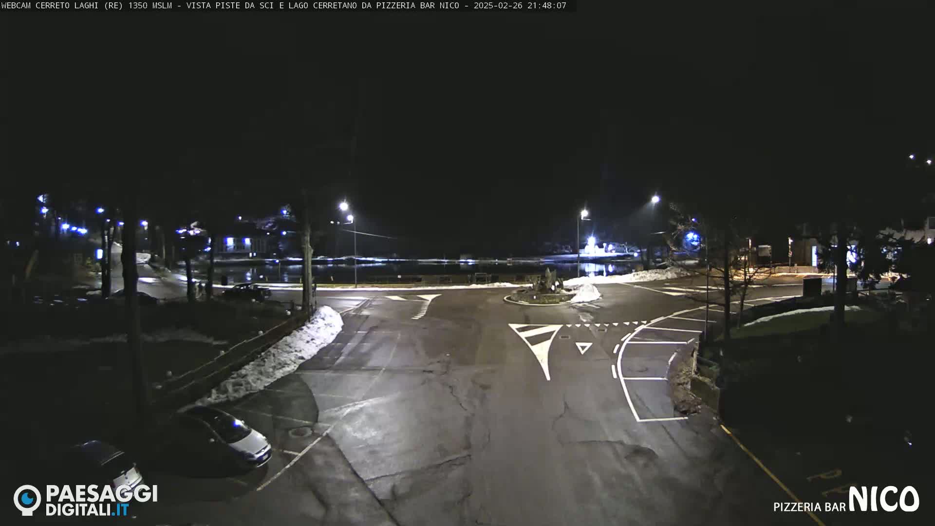 A nighttime view of a parking lot and roadway intersection with some snow on the ground near a body of water.