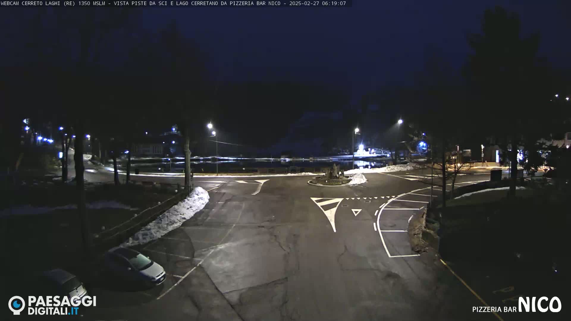A nighttime, snow-dusted parking lot and roadway intersect at a roundabout near a body of water under a dark sky.