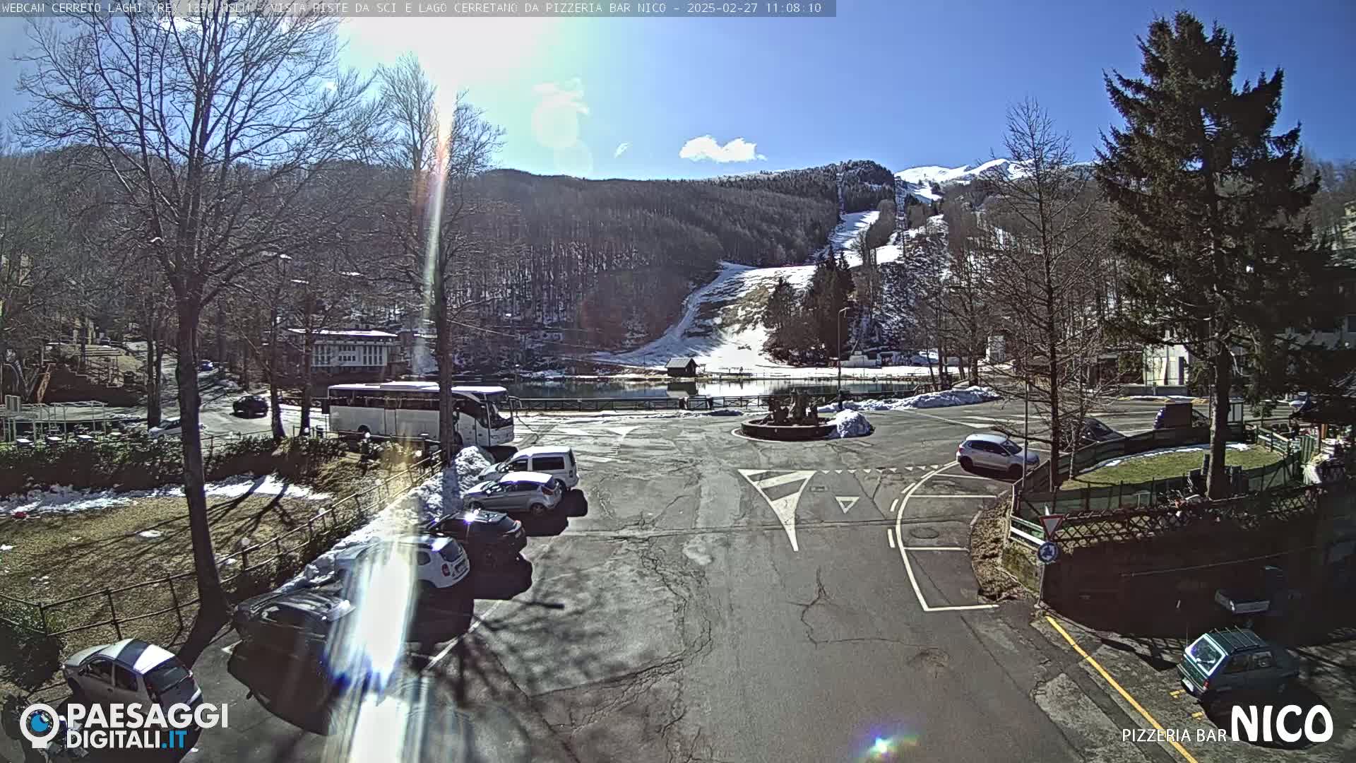A sunny parking lot with several cars and a bus, situated near a lake and snowy ski slopes in a mountainous area.