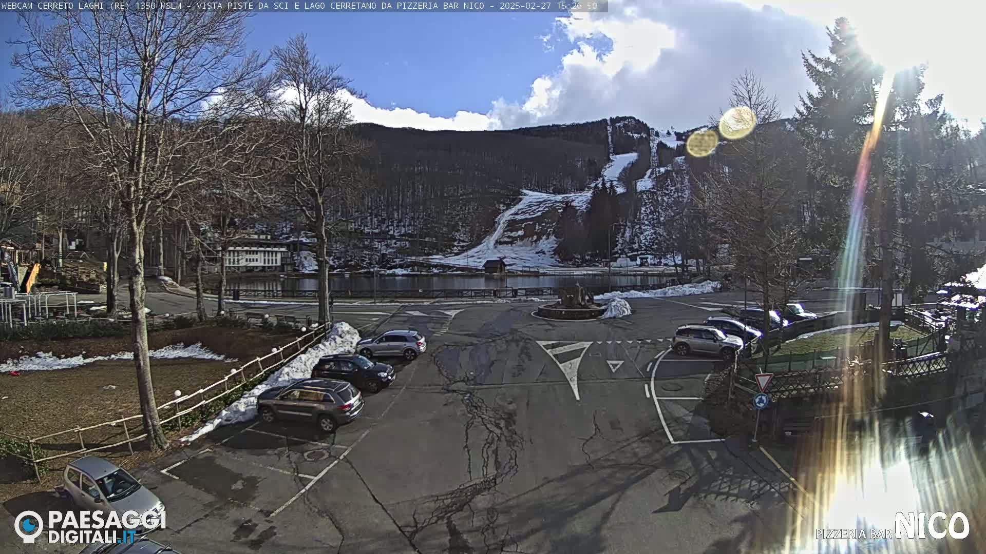 A sunny day reveals a parking lot with several cars, a small lake, and snow-dusted ski slopes on a hillside in the background.