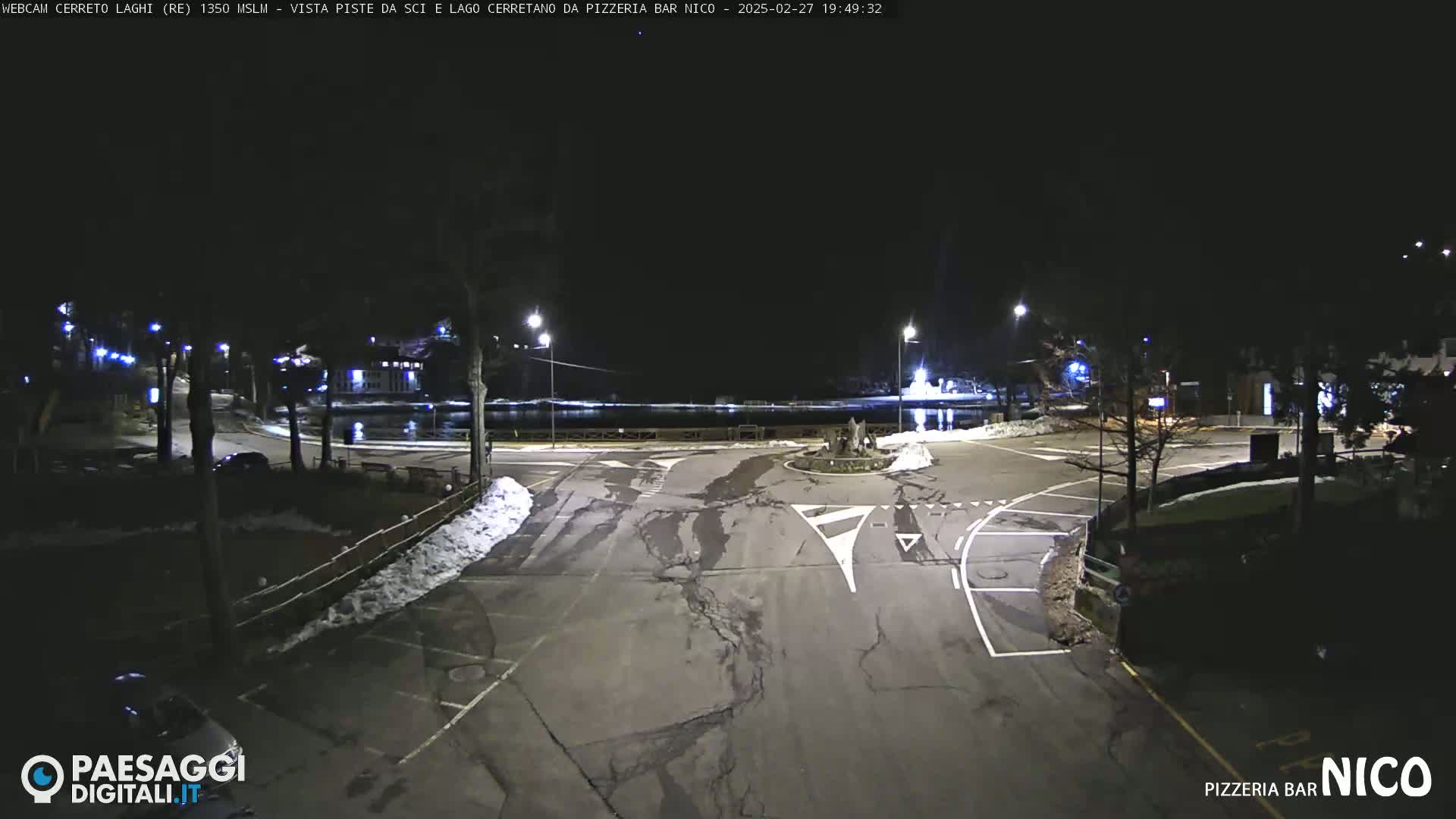 A nighttime view of a mostly empty parking lot and roadway next to a body of water, with some snow on the ground.