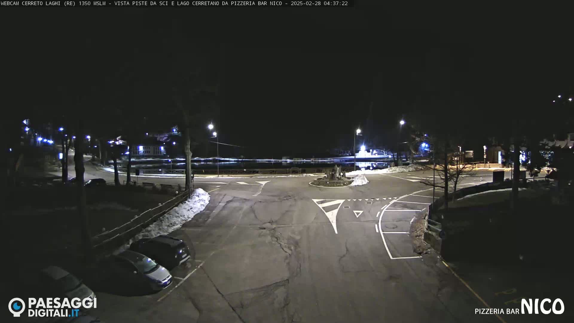 A nighttime view of a mostly empty parking lot and roadway intersection, illuminated by streetlights, with some snow on the ground.