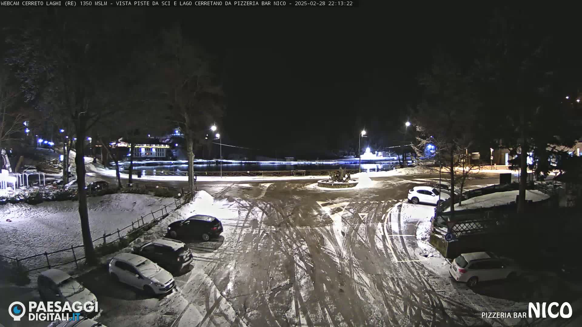 A snow-covered parking lot at night, with several cars parked and a body of water visible in the background under nighttime lighting conditions.