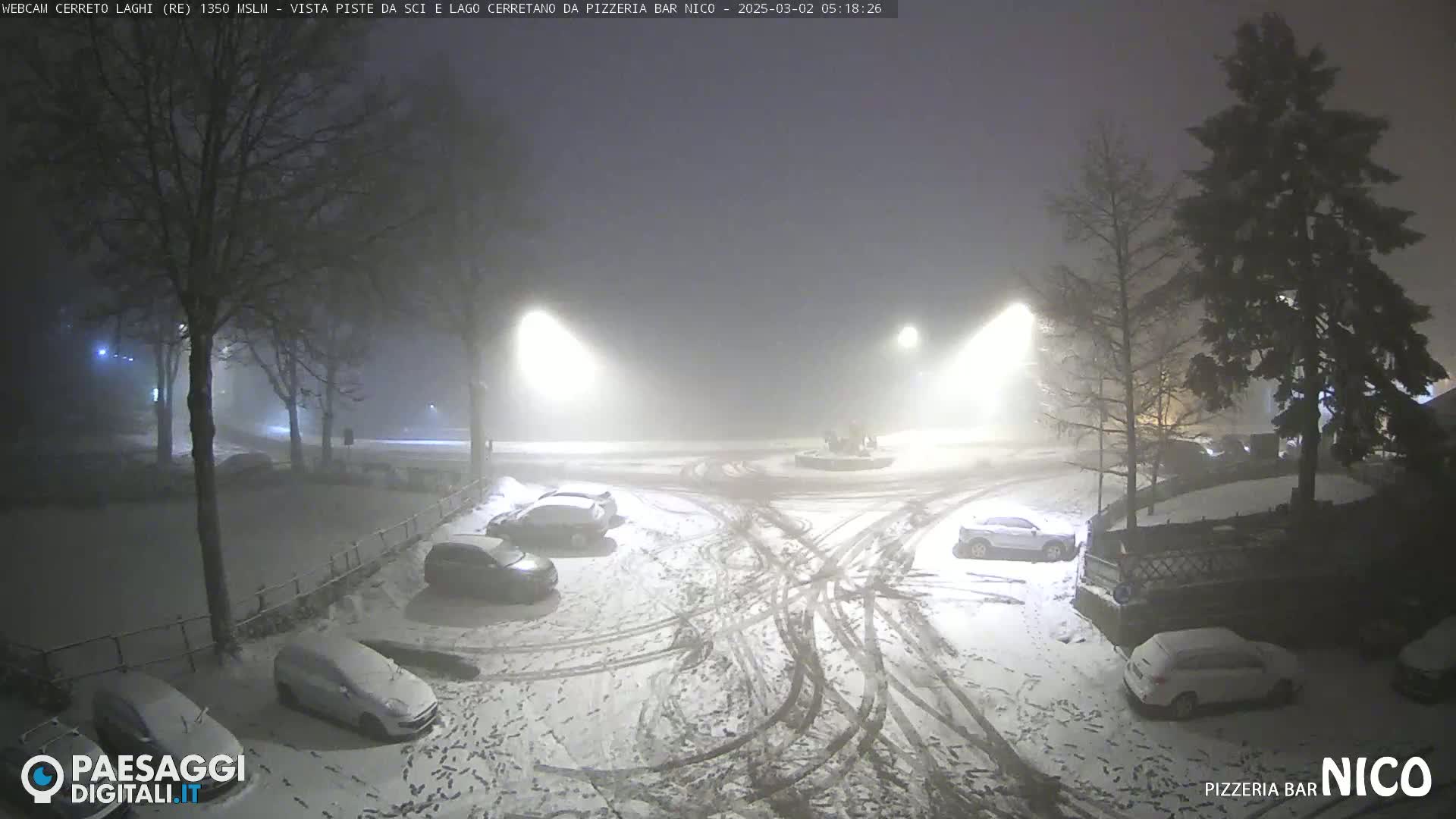 A snow-covered parking lot at night, dimly lit by streetlights in foggy conditions, with several cars parked and tire tracks visible in the snow.