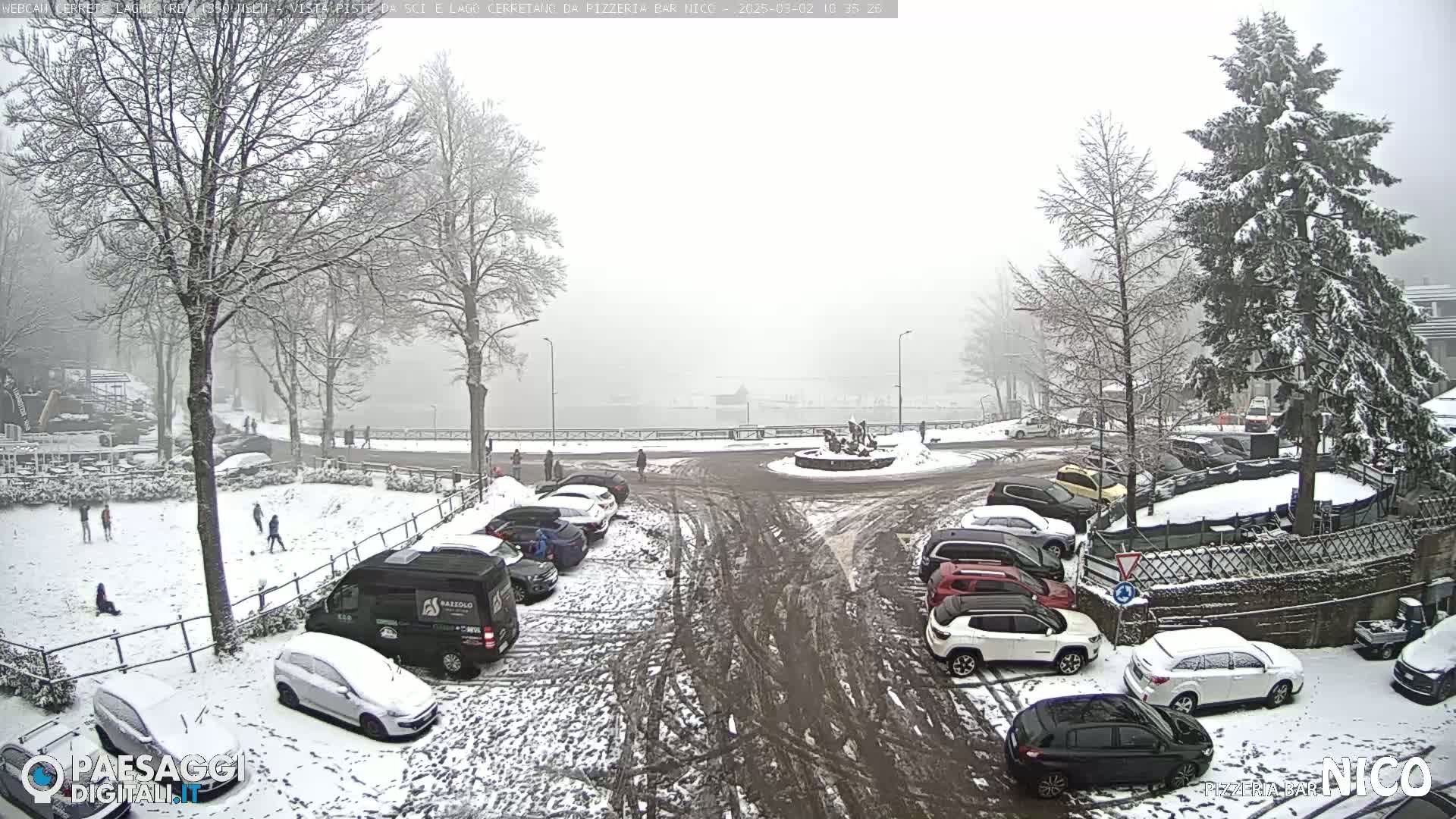 A snow-covered parking lot and surrounding area with several cars parked, some people walking, and a few children playing in the snow, all under a foggy, snowy sky.