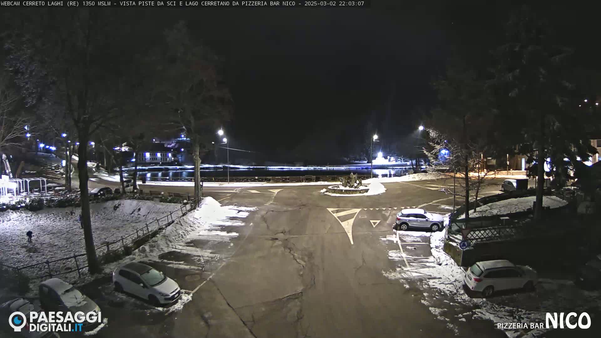 A snow-covered parking lot at night, with several cars parked and a body of water visible in the distance.
