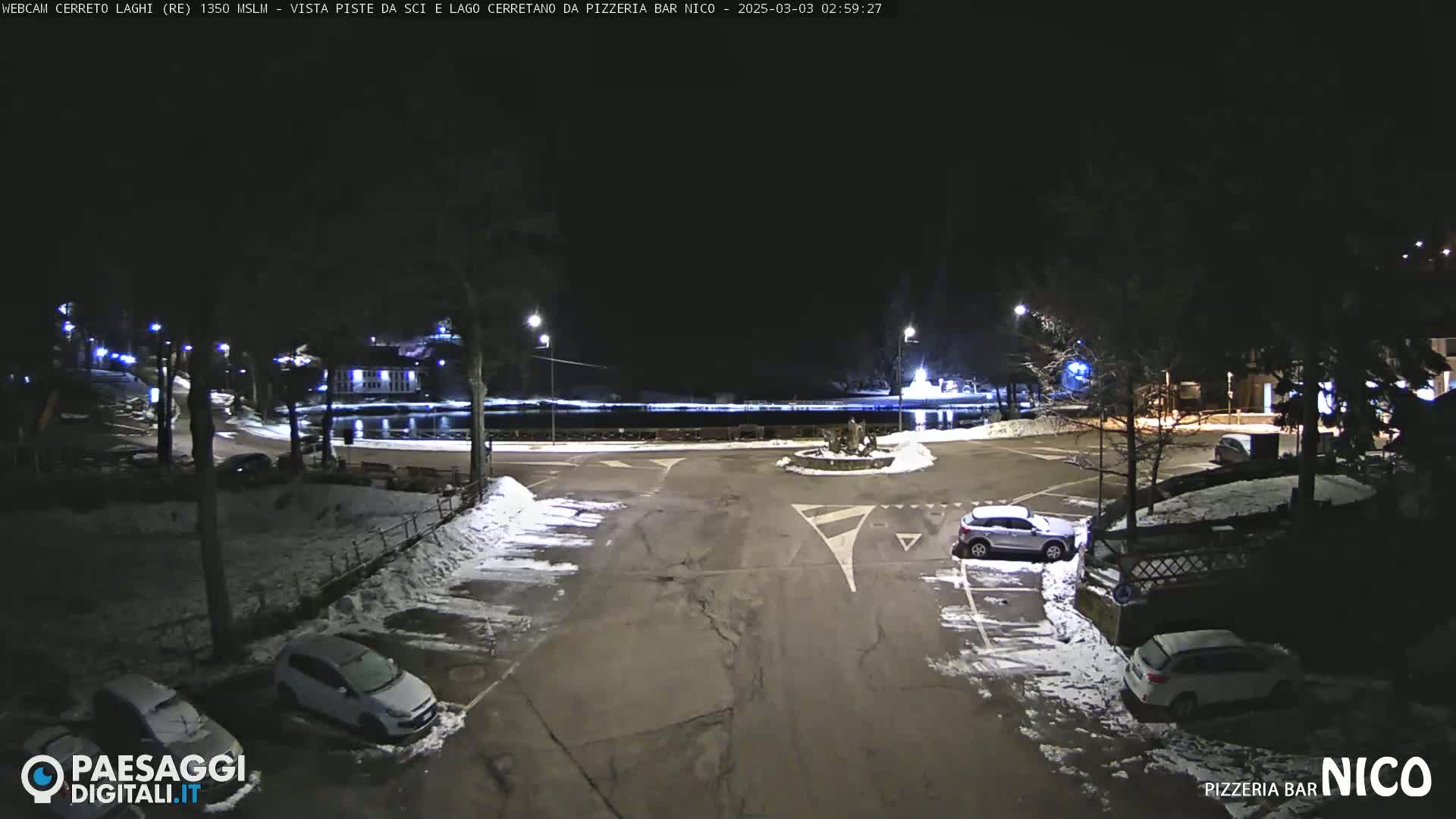 A nighttime, snow-covered parking lot and roundabout surround a calm body of water, illuminated by streetlights.