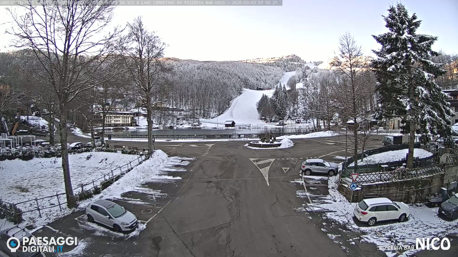 A snow-covered parking lot with several cars and a view of a ski slope in the background under a partly cloudy sky.