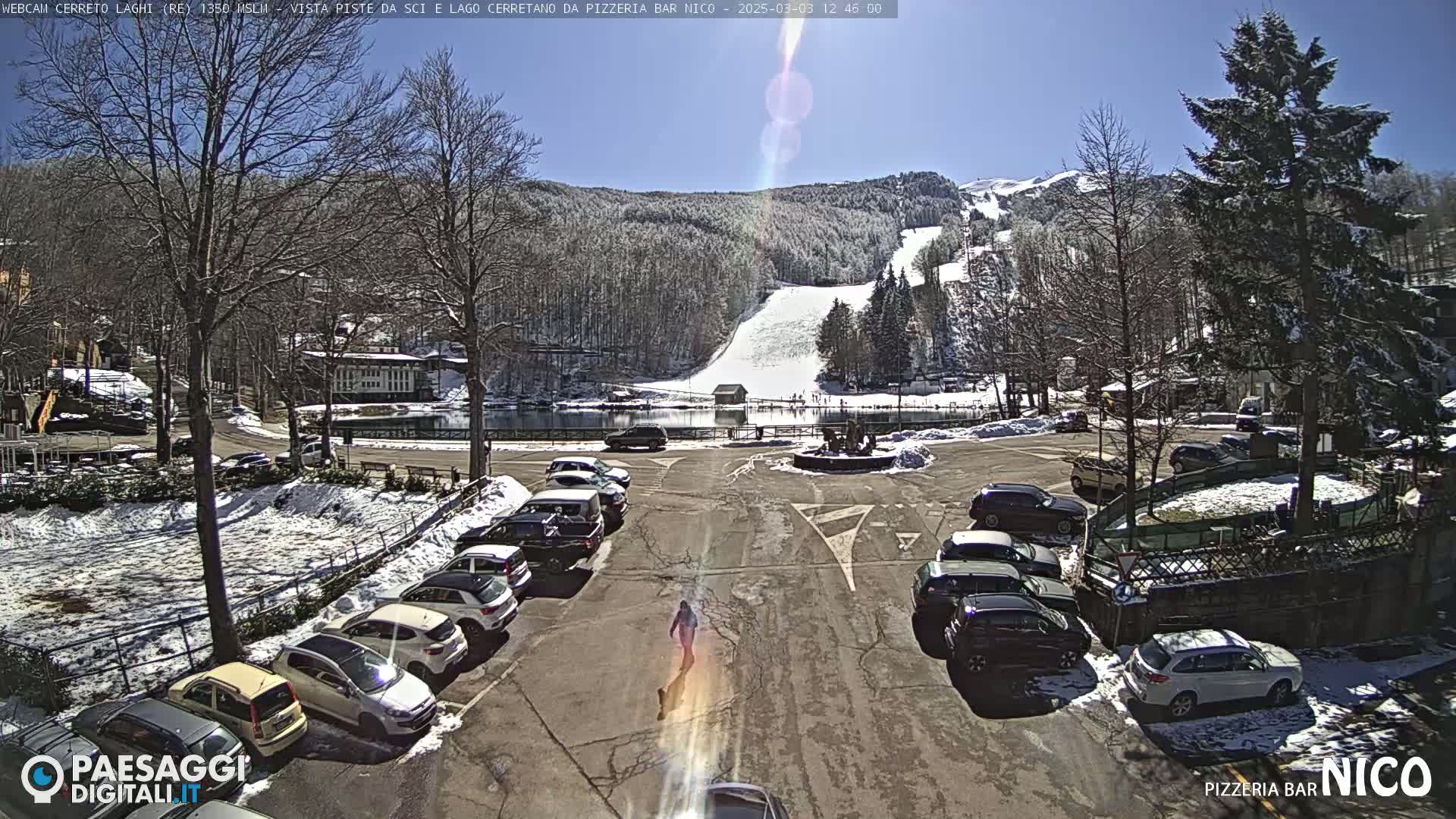 A sunny parking lot with cars and snow on the ground is situated near a lake and ski slope in a mountainous area.