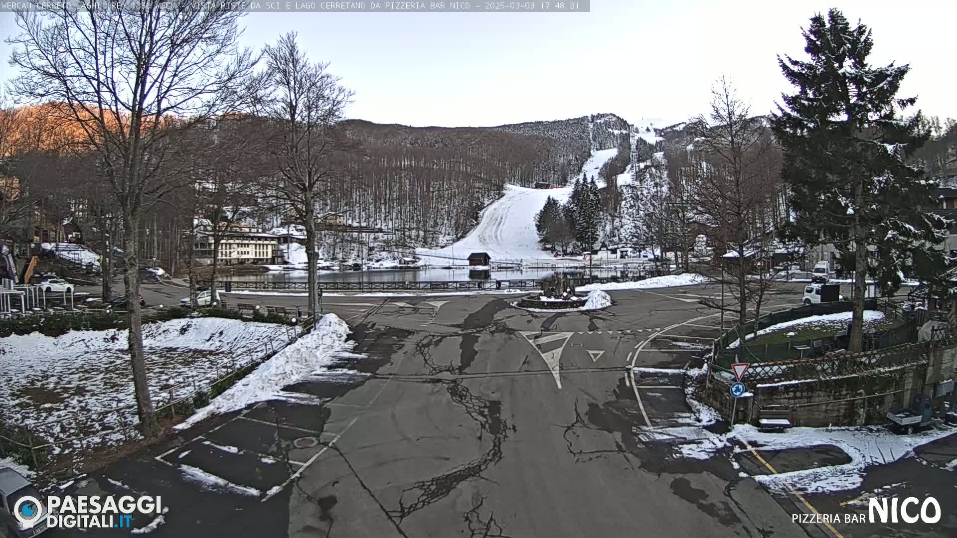 A snow-covered parking lot and road are situated in front of a small town nestled at the base of a snow-dusted mountain with ski slopes visible.